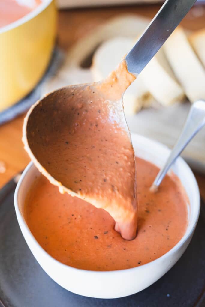 A large silver ladle pours tomato soup into a white ceramic bowl.