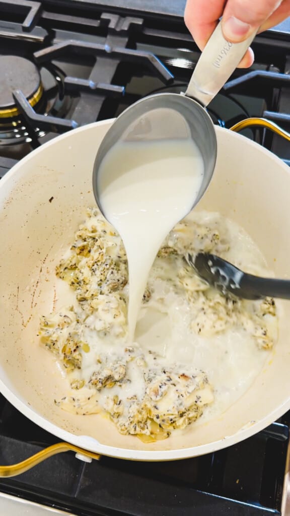 Milk from a measuring cup is poured into a pot of onions, cream cheese and seasonings sitting on top of a stovetop.