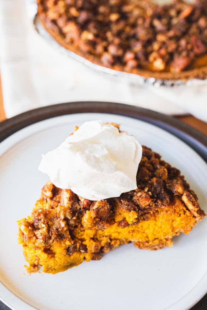 A slice of pecan streusel pumpkin pie sits on a white plate with a pie tin behind.
