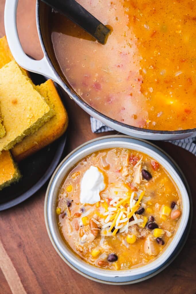 Bowl topped with cheese and sour cream sits beside a large pot of taco soup and plate of cornbread.