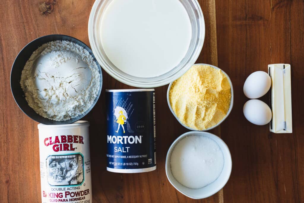 Ingredients for Sweet Homemade Corn Bread sit on a wooden countertop, portioned in individual bowls.