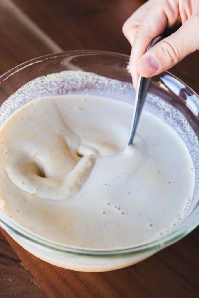 Milk and cornmeal are mixed together in a large glass bowl. A hand stirs the mixture with a silver spoon.