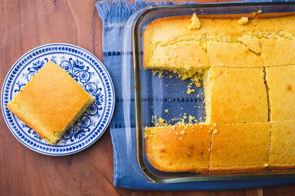 Slice of cornbread sits on a blue and white plate while beside a pan of sliced cornbread.