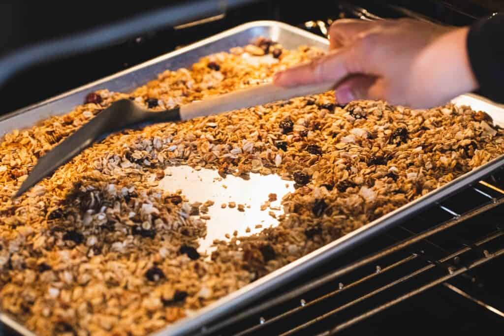 A spatula moves the granola cereal across a baking sheet in the oven.