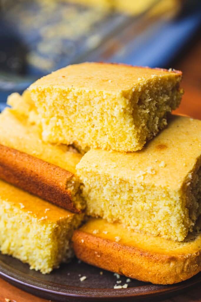 Squares of cornbread sit stacked on top of one another on a dark brown plate.