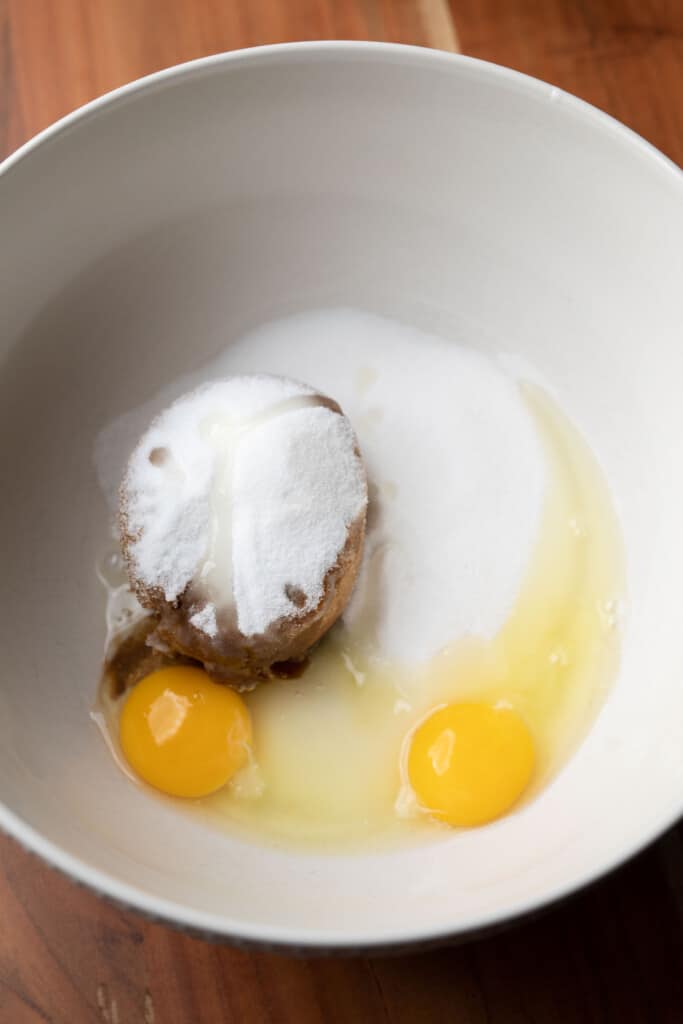 White and brown sugar sit in a white bowl with two eggs and vanilla.