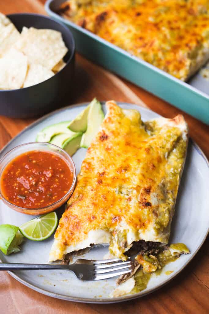 Two beef enchiladas verde sit on a plate with sliced avocados and a bowl of salsa with a fork.