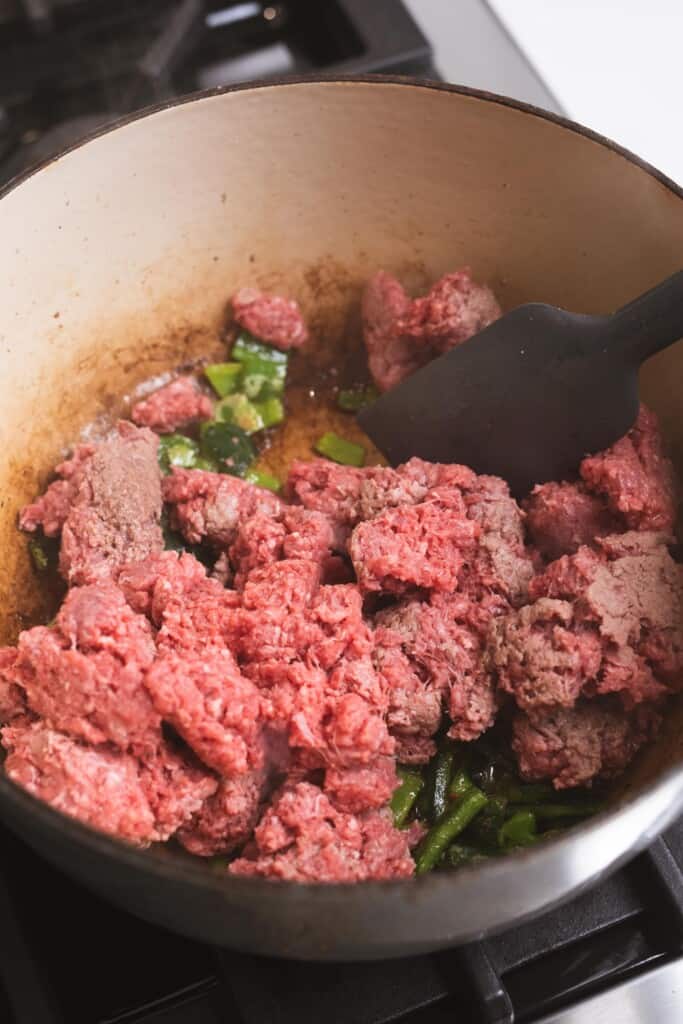 Raw ground beef and sauted peppers sit in a large pot on the stove.