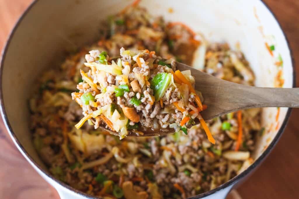 A wooden spoon stirs egg roll in a bowl filling, mixing everything together over a large white pot.