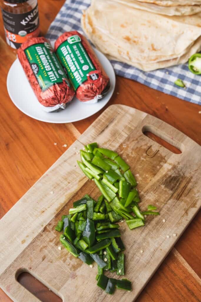 Poblano peppers sit sliced on a wooden cutting board with other ingredients sitting behind.