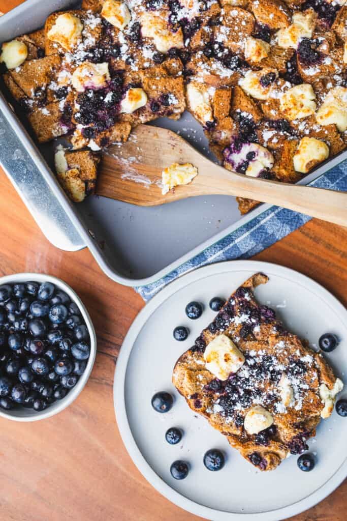 Golden brown French Toast Bake sits in a pan beside a serving of the casserole on a small white plate.