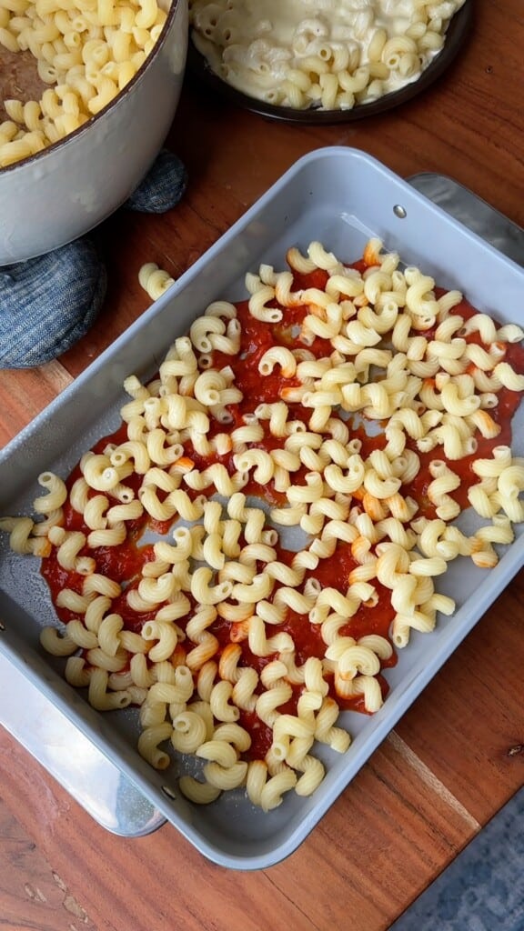 Marinara and curly noodles sit in the bottom of a pan.