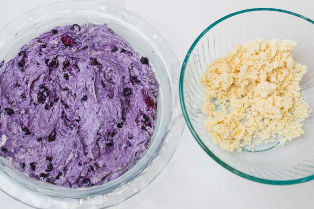 Purple muffin batter sits in a glass bowl beside a bowl of prepared streusel topping.