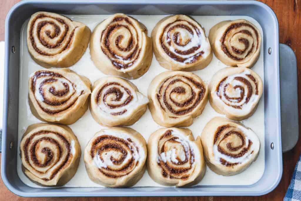 Warm heavy cream is poured over the top of the baking dish so that it fills the bottom between the spaces in between each cinnamon roll spiral.