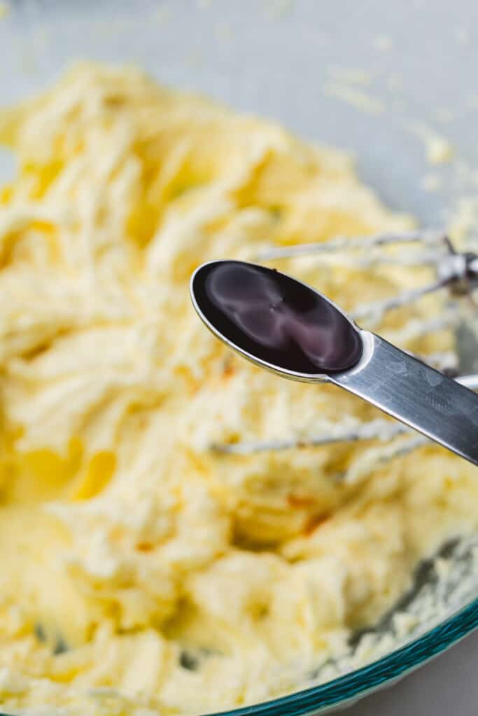 A measuring spoon full of vanilla sits over a bowl of creamed sugar and butter.