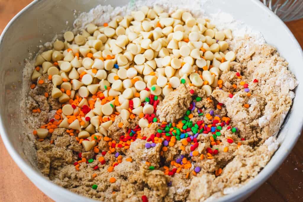 White chocolate chips and rainbow confetti sprinkles are added to the bowl of cookie dough ready to be folded in.