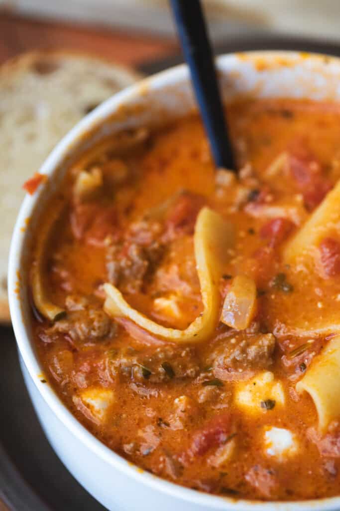 Bowl of lasagna soup sits on a plate with slices of warm bread.