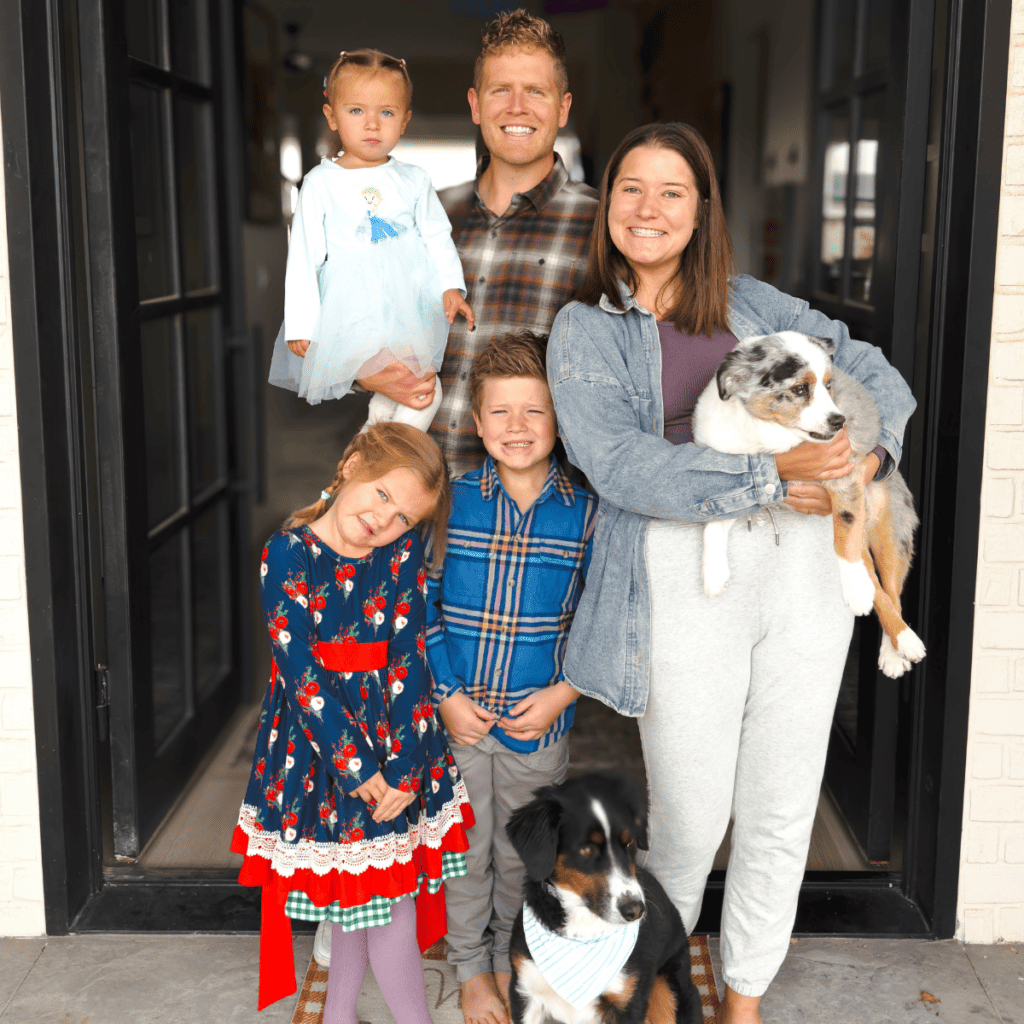 The Seely Family stands in front of an open door way. Dallin holds Violet while James and George stand in front of him. Ashley holds Luna in her arms and Dash sits quietly at their feet.