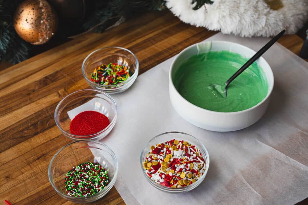 White chocolate died green sits in a bowl surrounded by 4 other small bowls of sprinkles.