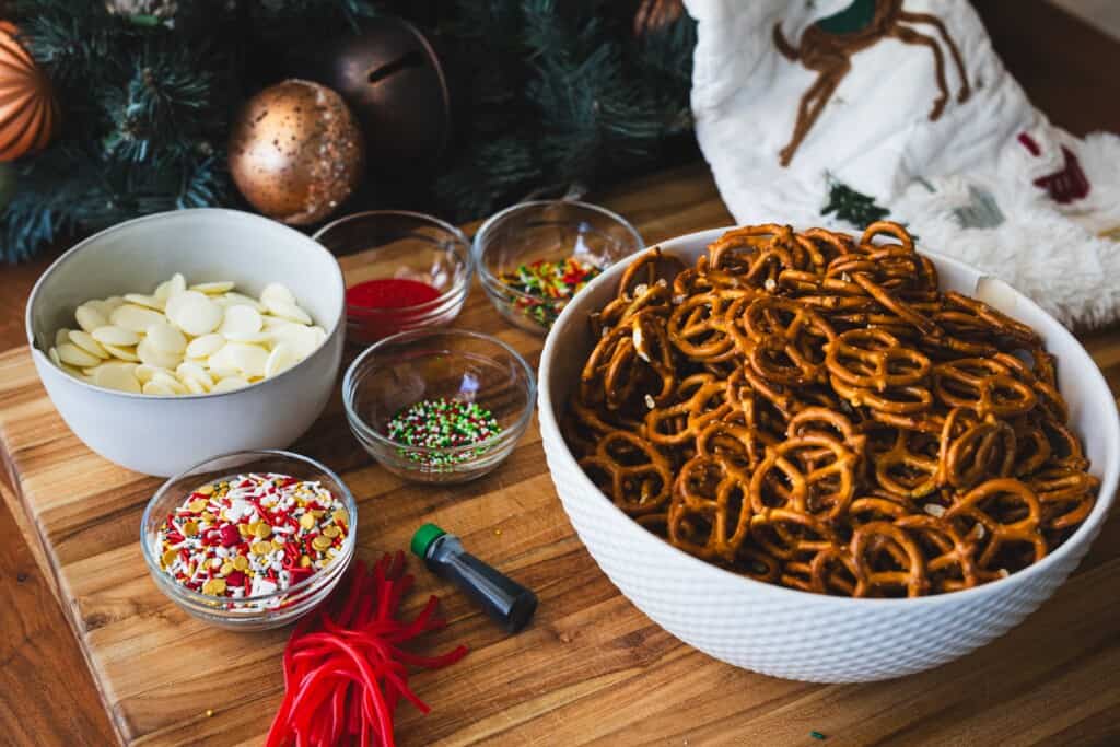 Ingredients for pretzel wreaths sit in bowls. White chocolate wafers, 4 kinds of sprinkles, green food coloring, pretzels, red licorice strings.