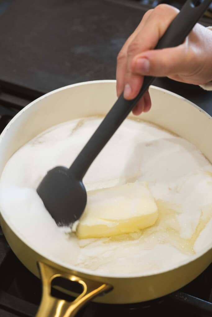 A black spatula is held over a pot with corn syrup, stick butter and sugar.