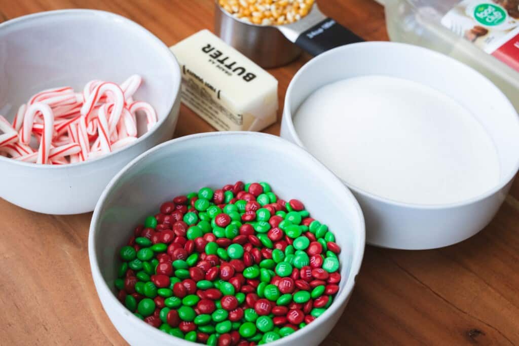Green and red mini m&ms sit in a white bowl beside a bowl of miniature candy canes and the other ingredients.