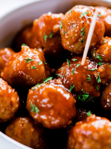 Honey garlic meatballs sit in a white bowl with a toothpick inserted for snacking.
