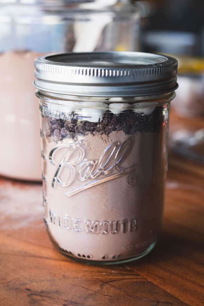 Small mason jar sits on wooden counter filled with hot chocolate mixture and topped off with mini chocolate chips and marshmallows.