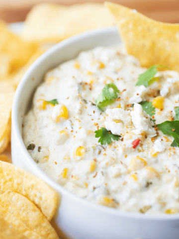 White bowl of Mexican Street Corn dip sits surrounded by crisp tortilla chips with a chip dipped in the bowl.