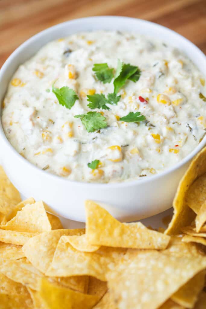 White bowl of Mexican Street Corn dip sits surrounded by crisp tortilla chips.