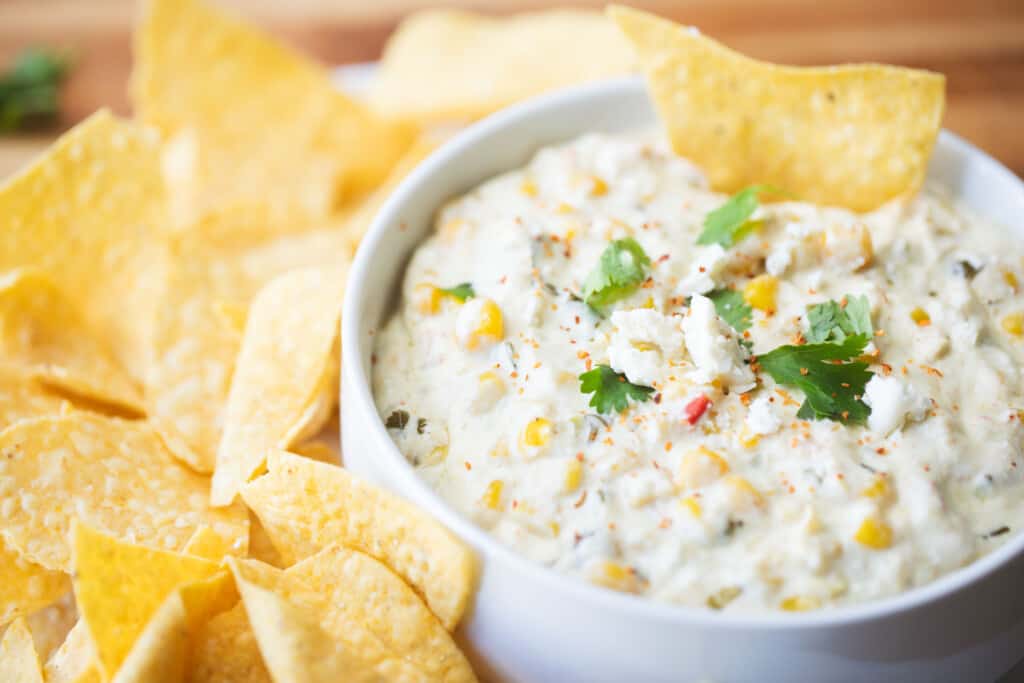 White bowl of Mexican Street Corn dip sits surrounded by crisp tortilla chips on a super bowl food table.