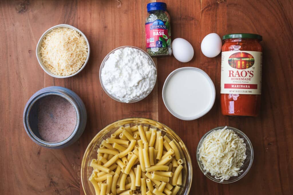 Ingredients for baked ziti sit in individual bowls on a wooden counter top.