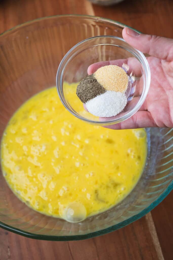 A hand holds a small glass bowl with three different seasonings over a larger bowl of raw scrambled eggs.