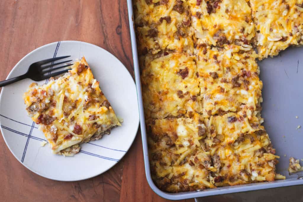 A served piece of Amish Breakfast Casserole sits on a plate with a fork beside the remaining casserole in the baking dish.