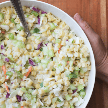 Two hands hold a bowl of creamy Coleslaw Pasta Salad sitting in a white serving bowl with metal spoon.