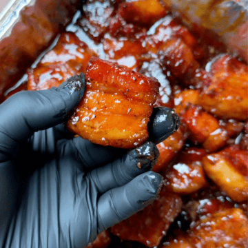 A black gloved hand holds a cut of pork belly burnt ends over a full pan of meat.