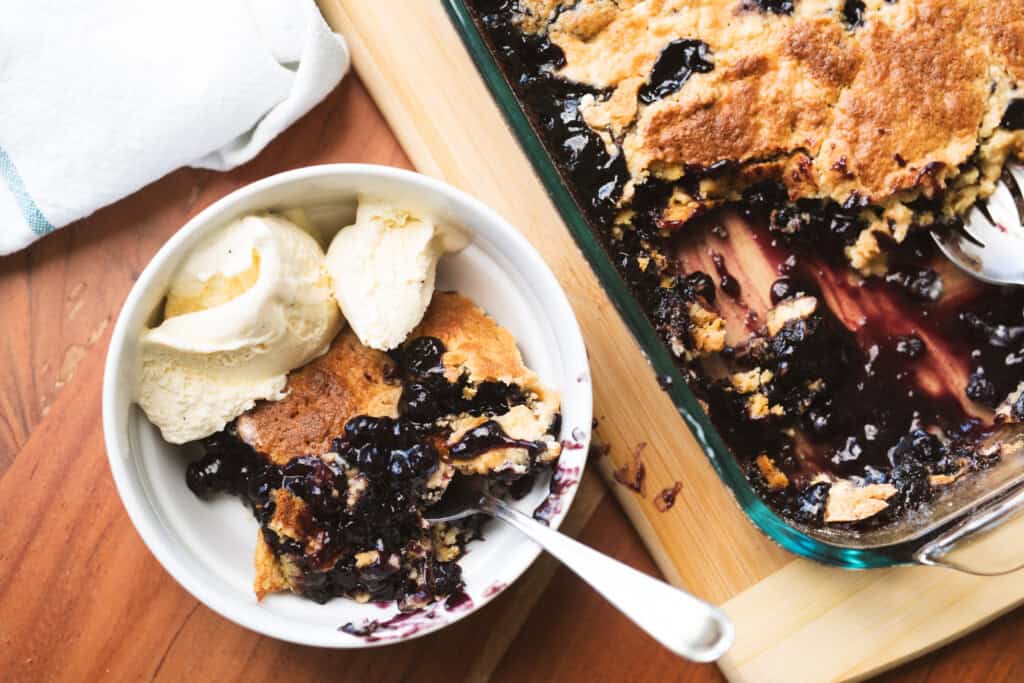 Bowl of blueberry cobbler is served a la mode next to the casserole dish.