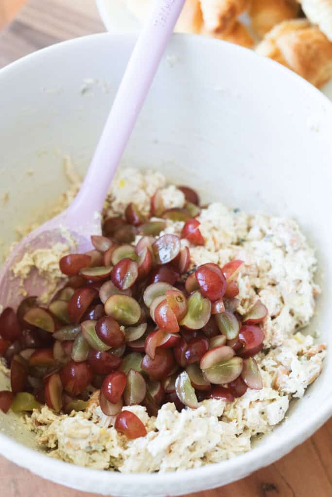 Grape halves are added to a large white mixing bowl with shredded chicken and seasonings.