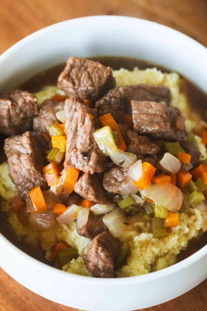 Serving of tender beef stew is served over a bed of mashed potatoes in a white bowl.