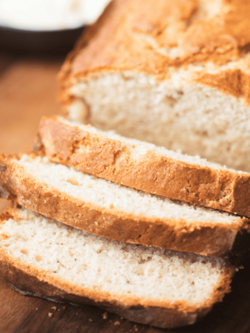 3 slices of bread sit on a wooden cutting board in front of a loaf of Cream Cheese Banana bread.