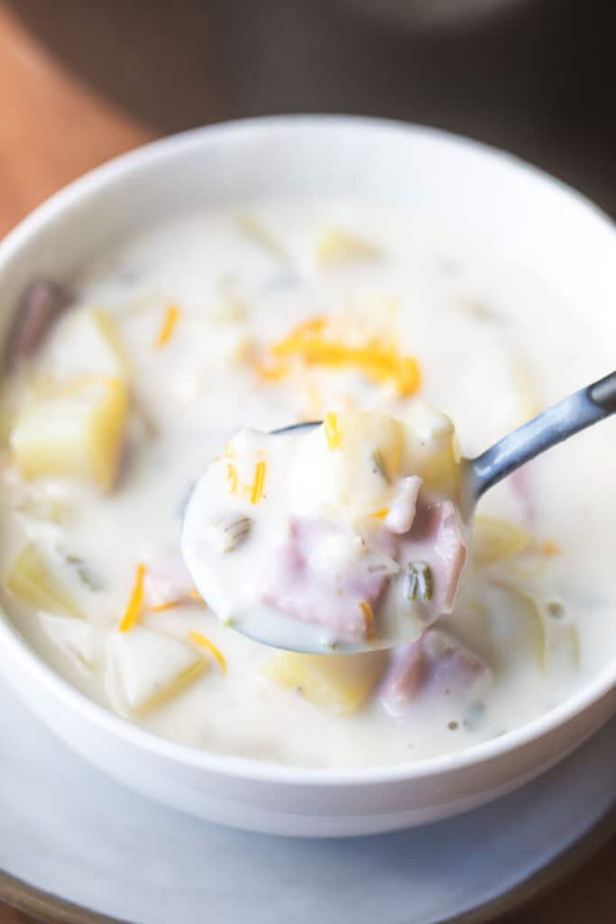 Spoonful of Ham and Potato soup is held over white ceramic bowl on a plate.