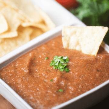 Fresh restaurant style salsa is served in a white ceramic dish beside crisp tortilla chips with a single chip dipped in.