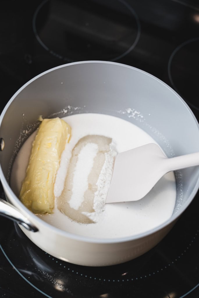 Coconut milk, stick butter, and sugar sit in a pot over the stove. A spatula stirs ingredients together.